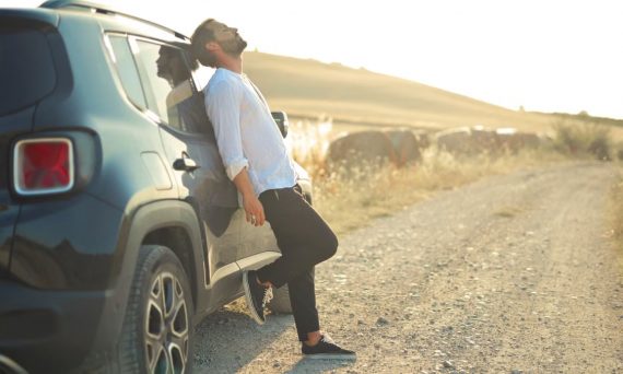 A man leans back on his SUV parked on a dirt road during a serene sunset, embodying relaxation and travel.